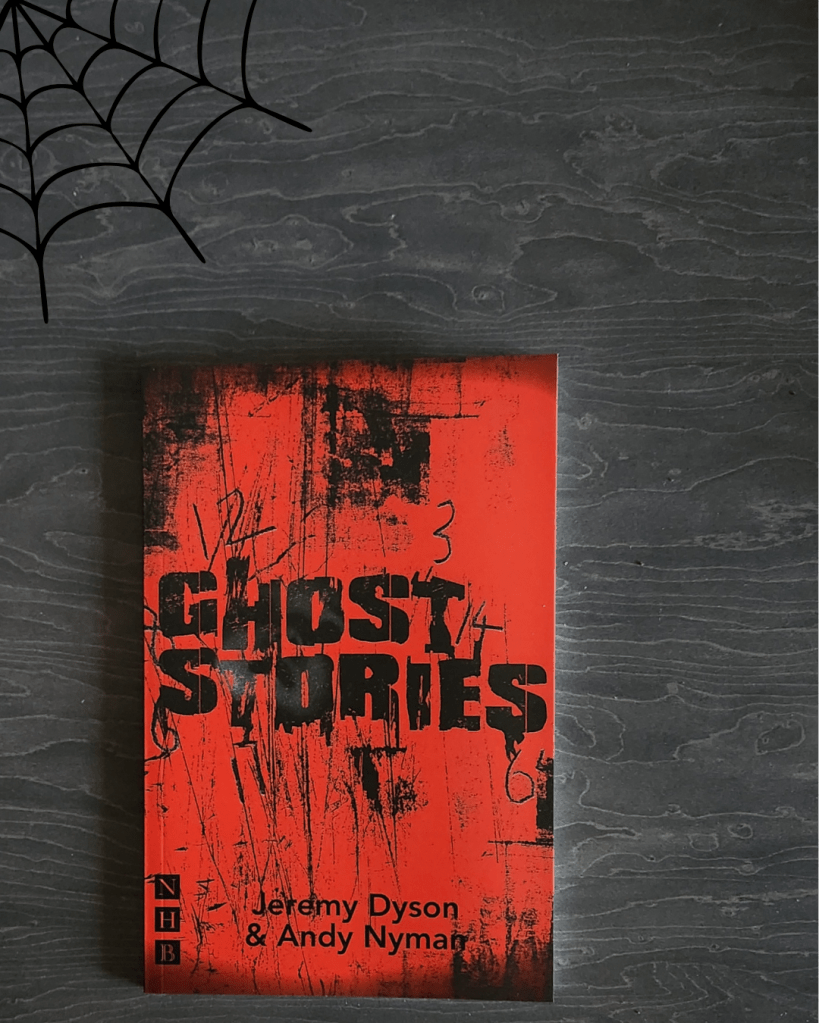 A dramatic flat lay featuring the play Ghost Stories by Jeremy Dyson and Andy Nyman. The book has a distressed red and black cover with rough textures and faint pencil markings. It rests on a dark wood surface with a simple black spider web graphic in the top left corner, adding a spooky Halloween touch.