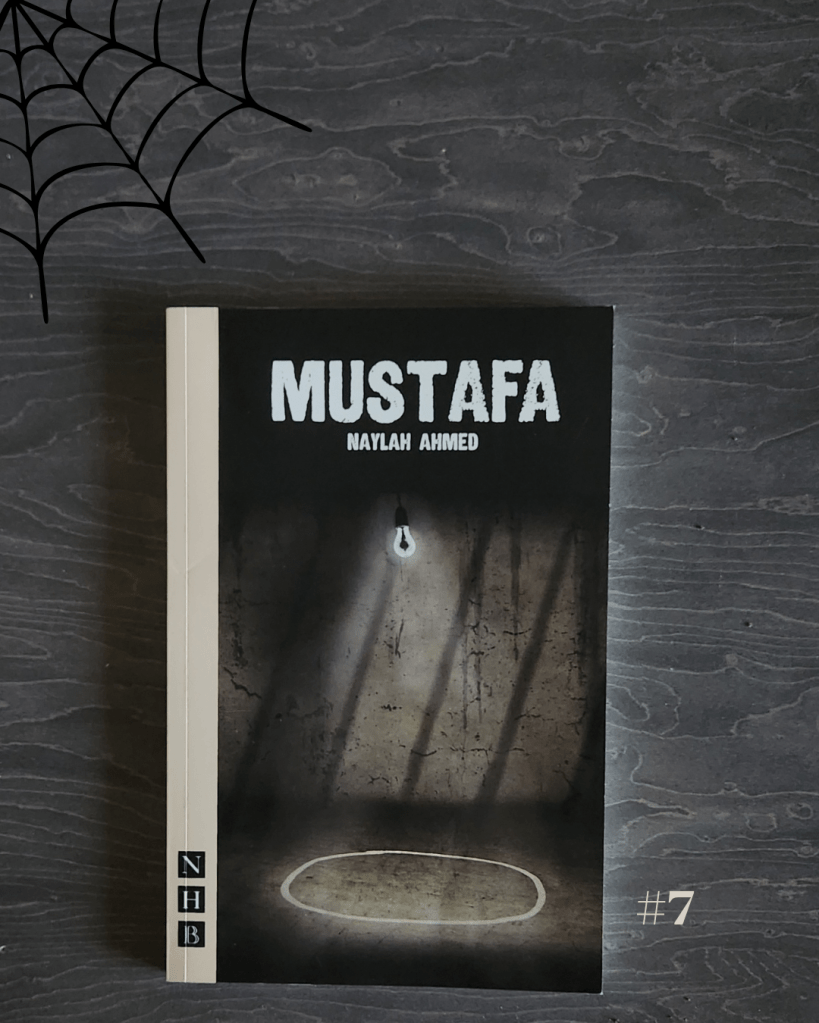 A flat lay of the play Mustafa by Naylah Ahmed, published by Nick Hern Books (NHB). The book cover shows a stark, dimly lit room with a single hanging light bulb casting shadows on a rough wall. A faint white chalk circle is drawn on the floor beneath the bulb, evoking a tense and eerie atmosphere. The cover rests on a dark wood surface with a black spider web graphic in the upper left corner and the number “#7” in light gray at the bottom right, maintaining a spooky, Halloween-inspired theme.