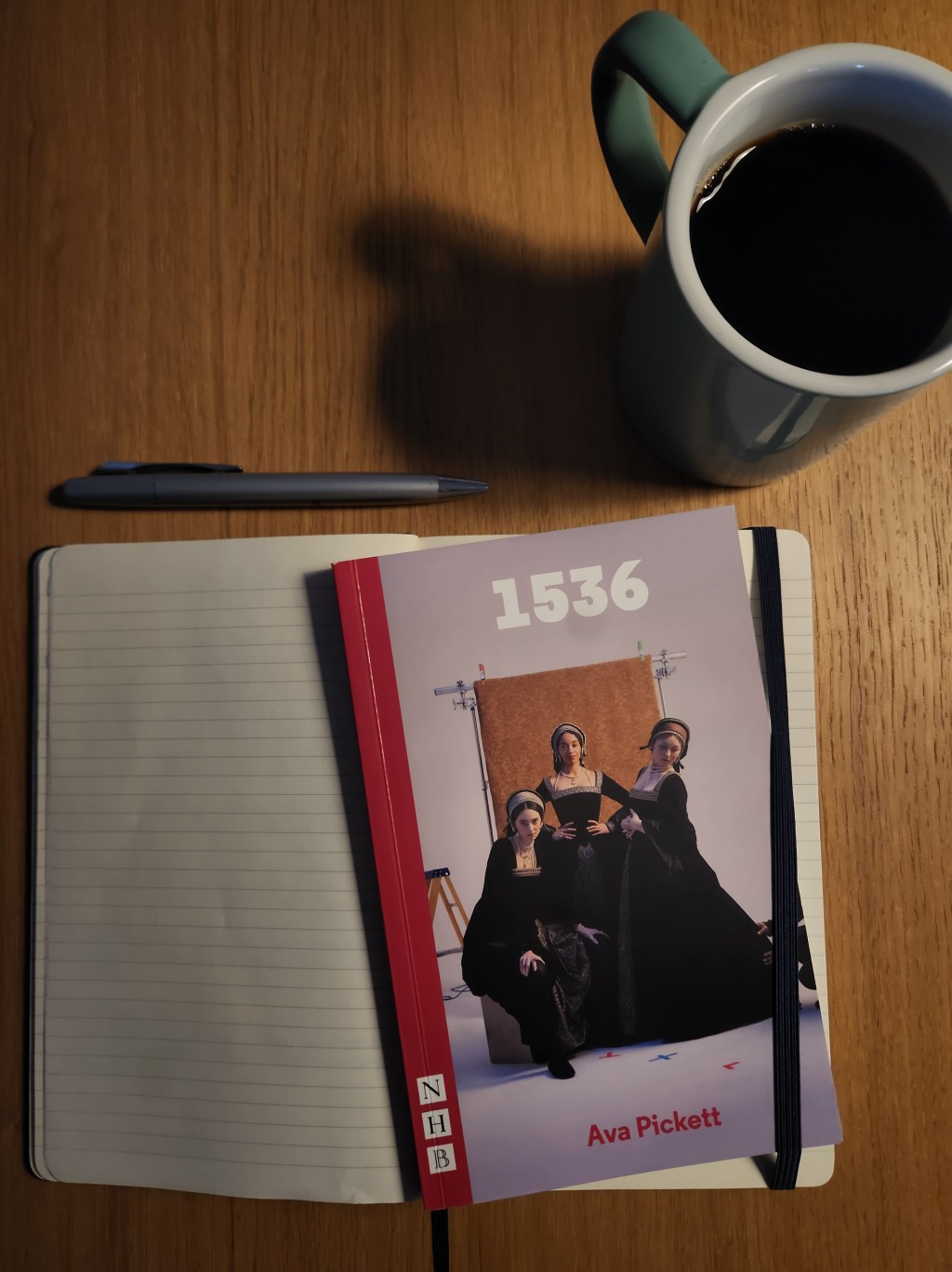A wooden desk with an open lined notebook, a gray pen resting above it, and a white mug filled with black coffee. On top of the notebook lies a book titled “1536” by Ava Pickett, featuring a cover image of three women in dark historical-style dresses posing in a studio setting.